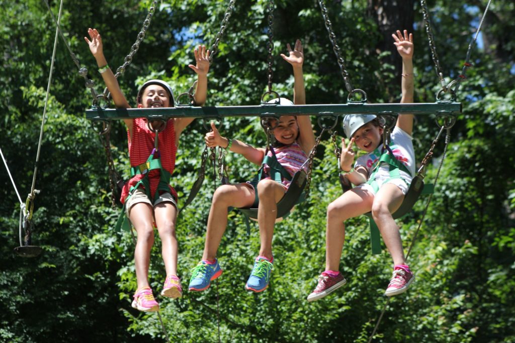 Young girls smile and wave while riding the 3-person swing.